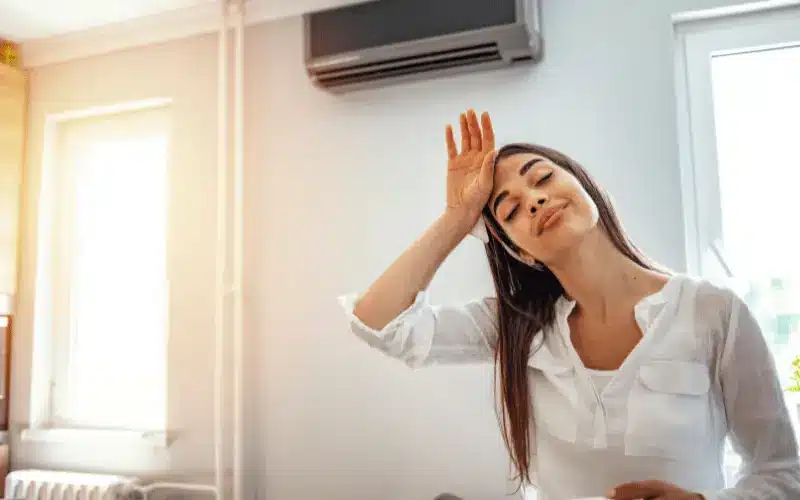 How To Prepare Air Conditioner for Summer 2 A woman wipes her forehead, looking uncomfortable from the heat in a room with an air conditioner.